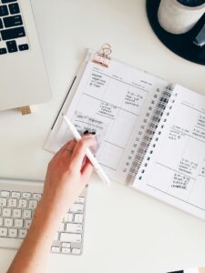 An open book lying on a desk next to a keyboard, with a person holding a pen above the book as if taking notes. If you want the alt text to emphasize something specific (like the type of book or the setting), feel free to provide more details